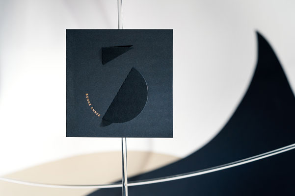 Cette photo représente la carte de vœux haut de gamme Calder, de la collection Intaglio 2023, sur une carte Bleu Savile, avec le détail de la gravure Cuivre et de ses découpes, et mise en scène dans un petit cirque de papier.